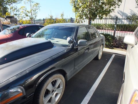 2003 Ford Crown Victoria - Dark Panther sedan shown side-on with alloy wheels, fence and tan building behind in a parking lot.