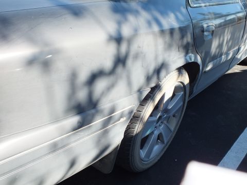 2003 Ford Crown Victoria - Front fender and alloy wheel of a dark sedan dappled with tree shadows.