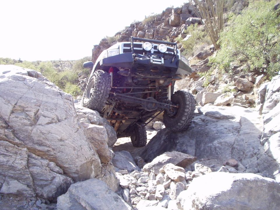 Lifted 4x4 crawling over desert boulders, underside showing solid axle, red shocks and winch bumper