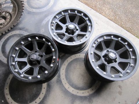 2008 Jeep Wrangler - Three black alloy wheels are displayed on a concrete surface, with a motorcycle tire in the background.