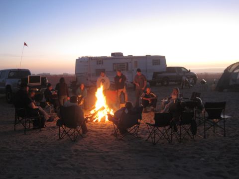 1989 Ford Bronco - A group of people enjoy a campfire on the beach as the sun sets, with camping equipment in the background.