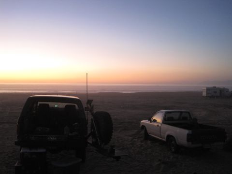 1989 Ford Bronco - Two trucks and RVs are parked on a beach as the sun sets over the ocean.