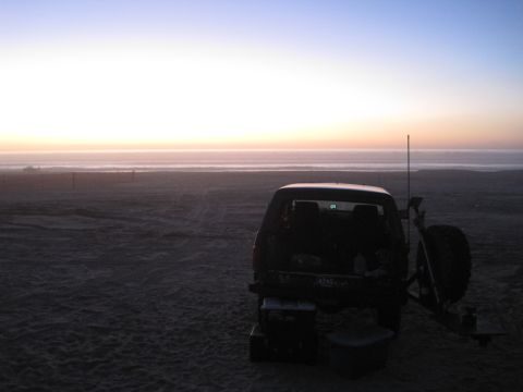 1989 Ford Bronco - A lone SUV sits on the sand as the sun sets over the ocean, casting a warm glow on the horizon.