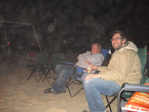 1989 Ford Bronco - Two men relax in camping chairs on a sandy area at night, with a bag of barbecue chips nearby.