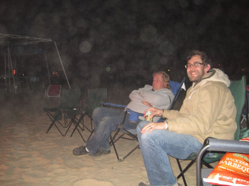Two men sit in camping chairs on sand at night, one holding a can, the other with arms crossed. A bag of barbecue chips is visible