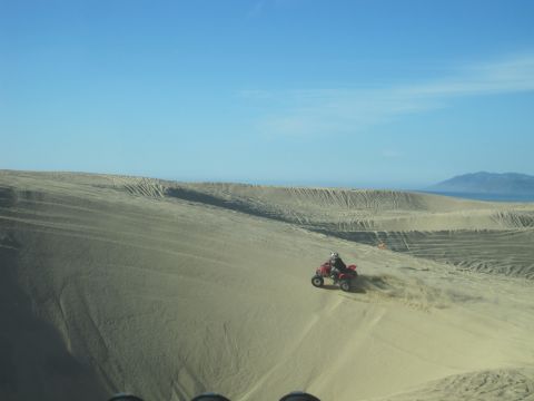1989 Ford Bronco - A red ATV navigates a sandy slope under a clear blue sky, kicking up dust as it goes.