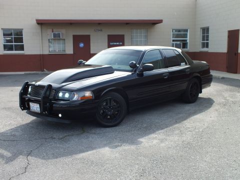 2003 Ford Crown Victoria - Black Crown Victoria sedan with push bar and black five-spoke wheels parked beside a tan building.