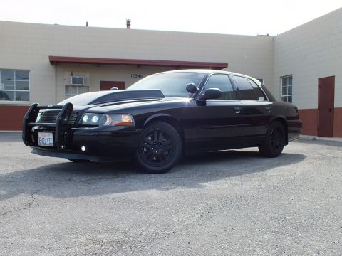 2003 Ford Crown Victoria - All-black full-size Ford sedan, low front quarter angle showing push bumper and tinted windows.