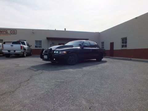 2003 Ford Crown Victoria - Murdered-out Ford sedan with push bumper parked next to a pickup outside an industrial building.