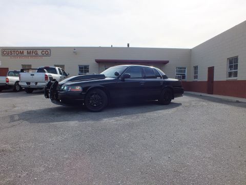 2003 Ford Crown Victoria - All-black full-size sedan front quarter, push bar visible, parked at an industrial site.