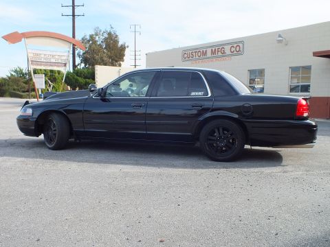 2003 Ford Crown Victoria - Side profile of murdered-out Ford sedan parked on pavement near a low industrial building.