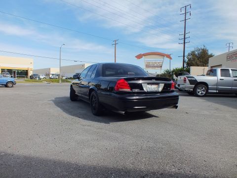2003 Ford Crown Victoria - Blacked-out Ford sedan from the rear, dual exhaust tips and black wheels, parked near trucks.
