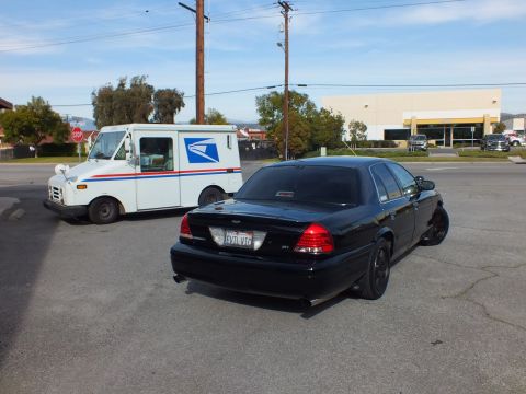 2003 Ford Crown Victoria - Murdered-out Ford sedan rear quarter next to a white postal delivery truck at an intersection.