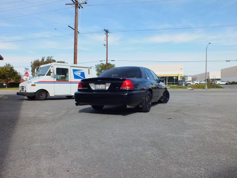 2003 Ford Crown Victoria - Blacked-out sedan rear quarter beside a white USPS truck under a partly cloudy sky.
