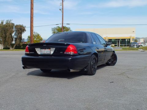 2003 Ford Crown Victoria - Murdered-out Ford sedan from the right rear with red taillights and black wheels in a lot.