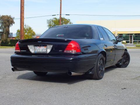 2003 Ford Crown Victoria - Murdered-out Ford sedan right rear view with Ford oval, P71 badge and black wheels.