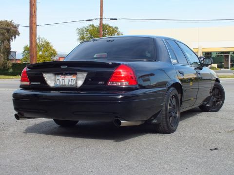2003 Ford Crown Victoria - Blacked-out full-size sedan from the right rear, dual exhaust tips and red taillights, sunny lot.