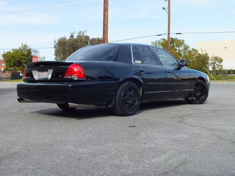 2003 Ford Crown Victoria - Rear quarter of a blacked-out sedan in a parking area, red taillights and black wheels visible