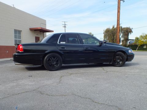 2003 Ford Crown Victoria - Full driver-side profile of a blacked-out sedan parked beside a tan industrial building