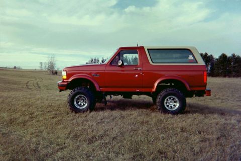 1995 Ford Bronco - A lifted red Ford Bronco with a tan removable top sits in a field under a cloudy sky.