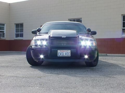2003 Ford Crown Victoria - Low stance Crown Vic head-on, projector and fog lights lit, EVIL VIC plate against a tan building backdrop.