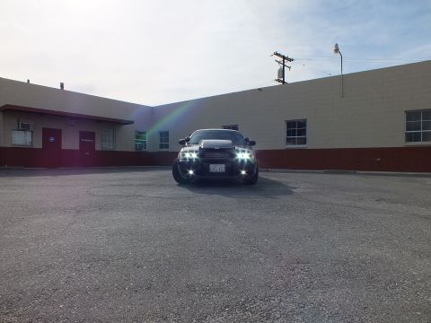 2003 Ford Crown Victoria - Wide shot of the black Crown Victoria across a paved lot, lights on, with a faint rainbow flare across the frame.