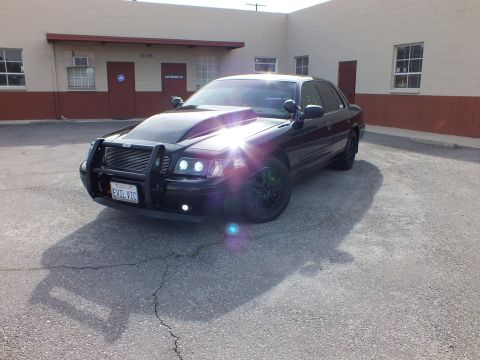 2003 Ford Crown Victoria - Three-quarter front of the murdered-out Crown Vic, push bar and grille guard, sun flaring off the fog light.