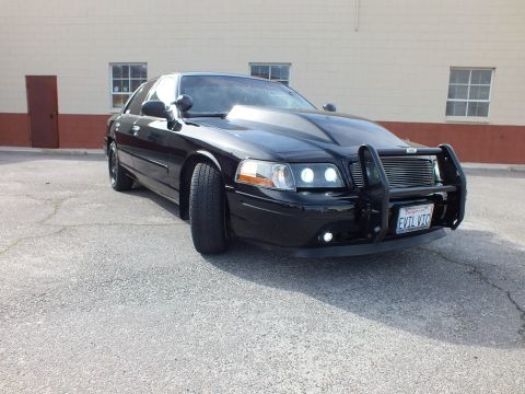 2003 Ford Crown Victoria - Glossy black Crown Vic from the front corner, grille guard and projector HIDs, EVIL VIC plate visible.