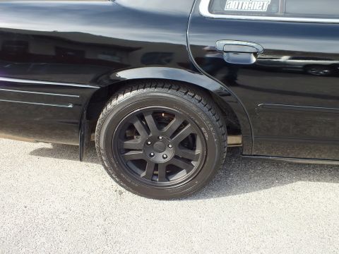 2003 Ford Crown Victoria - Rear quarter wheel detail: matte-black multi-spoke wheel with Ford cap, AUTO.NET decal on the glass above.