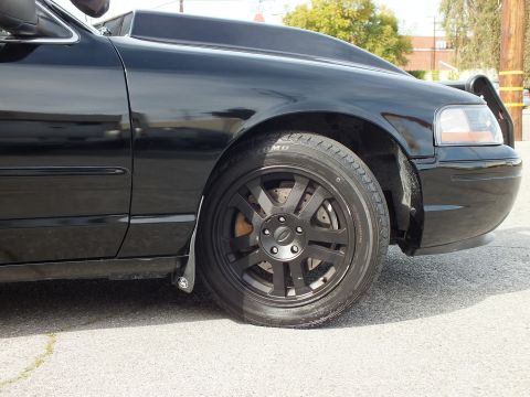 2003 Ford Crown Victoria - Front wheel close-up showing the matte-black multi-spoke rim, Ford cap and Toyo tire of the black sedan.