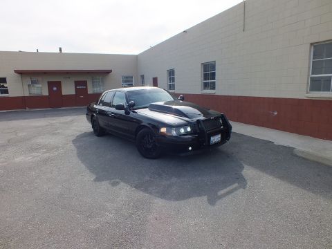 2003 Ford Crown Victoria - Distant front-quarter view of the murdered-out Crown Vic with grille guard against a two-tone building.