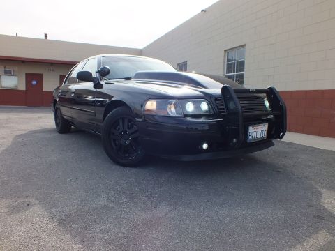 2003 Ford Crown Victoria - Front-quarter of the blacked-out Crown Vic, grille guard and projector HIDs lit, California plate in front.