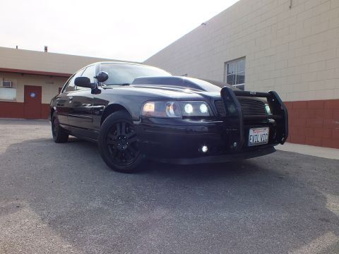 2003 Ford Crown Victoria - Front-corner shot of the murdered-out Interceptor, push bumper and twin HIDs against a tan wall.