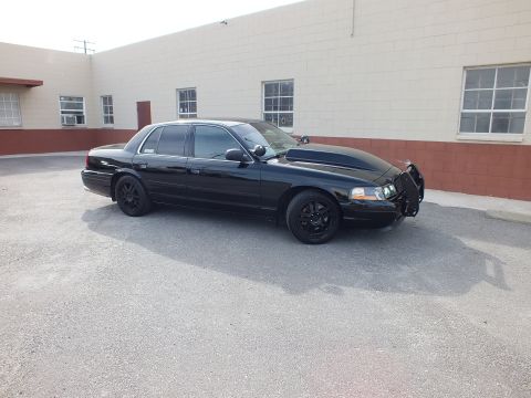 2003 Ford Crown Victoria - Side-front profile of the blacked-out Crown Vic, grille guard and black wheels, parked by a tan building.