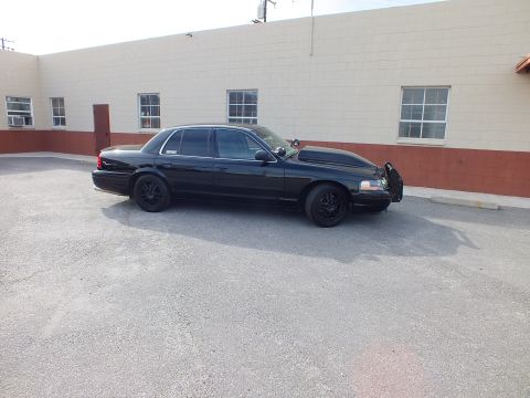 2003 Ford Crown Victoria - Murdered-out Crown Victoria shown from the passenger side on cracked asphalt beside a windowed building.