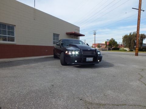 2003 Ford Crown Victoria - Black Crown Victoria with brush guard and lit headlights parked beside a gray building.