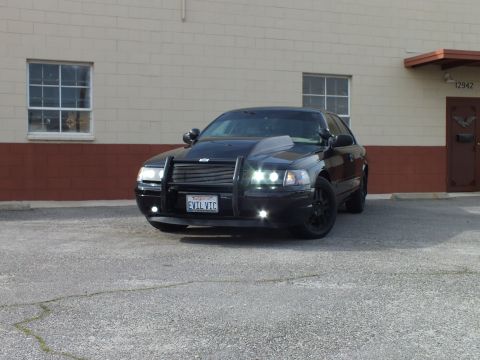 2003 Ford Crown Victoria - Black Crown Victoria with brush guard and fog lights parked by a windowed building.