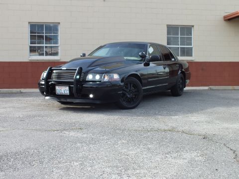 2003 Ford Crown Victoria - Black Crown Victoria with brush guard shown front-left beside a tan windowed building.