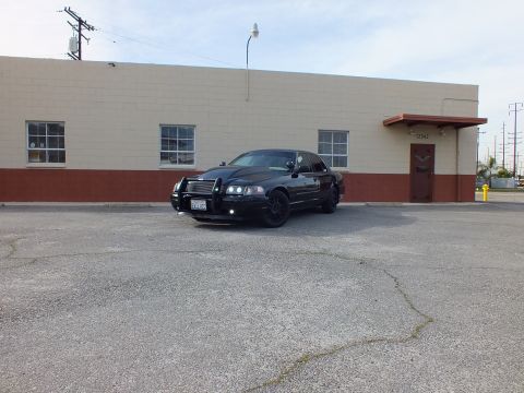 2003 Ford Crown Victoria - Black Crown Victoria with brush guard shot from across a cracked lot near a gray building.