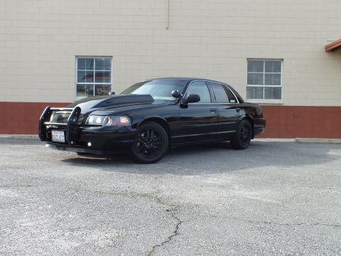 2003 Ford Crown Victoria - Murdered-out black Crown Victoria sedan, front quarter view, with brush guard and blacked-out alloy wheels on cracked asphalt.