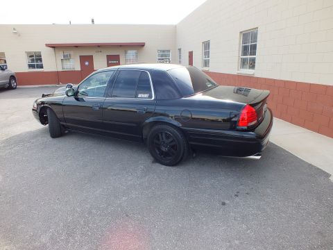 2003 Ford Crown Victoria - Rear driver-side view of a black Ford Crown Victoria, red taillights and black alloy wheels in a lot by tan buildings.