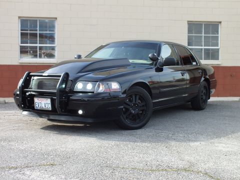 2003 Ford Crown Victoria - Black Crown Victoria, driver-front angle, brush guard and black wheels, headlights illuminated; plate EVIL VIC.