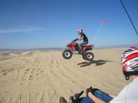 1989 Ford Bronco - A rider on a red ATV jumps over sand dunes under a clear blue sky.