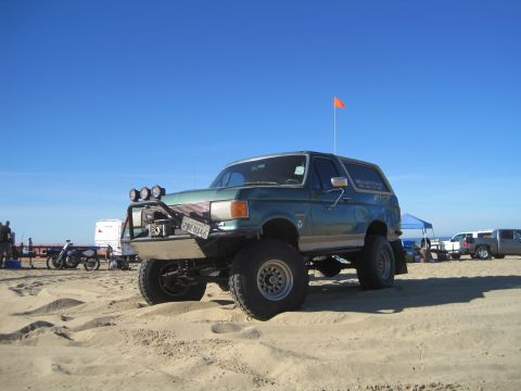 1989 Ford Bronco - A modified green Ford Bronco sits on a sandy beach under a clear blue sky, with other vehicles in the background.