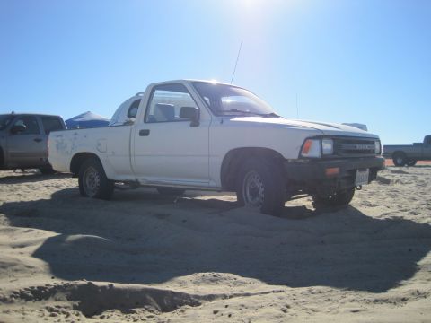 1989 Ford Bronco - White Toyota pickup truck parked on sand with other vehicles nearby under a clear blue sky.