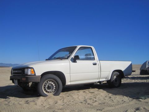 1989 Ford Bronco - White 1989 Toyota pickup truck with a flat front tire on a sandy surface under a clear blue sky.
