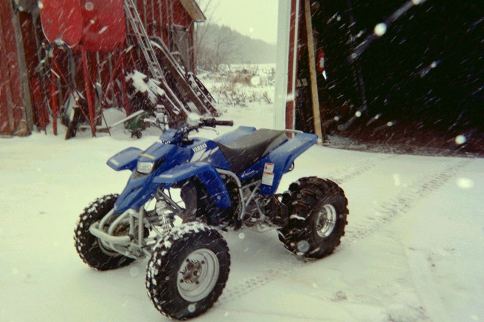 Blue Yamaha ATV parked in snow near a red barn with skis stacked