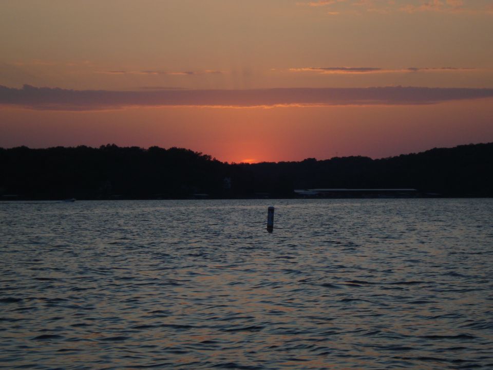 Sunset over a lake with a tree line in the background. A buoy is in the water