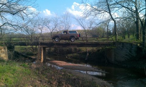 1987 Ford Bronco - A brown 1987 Ford Bronco traverses a weathered bridge over a shallow creek in a wooded area.