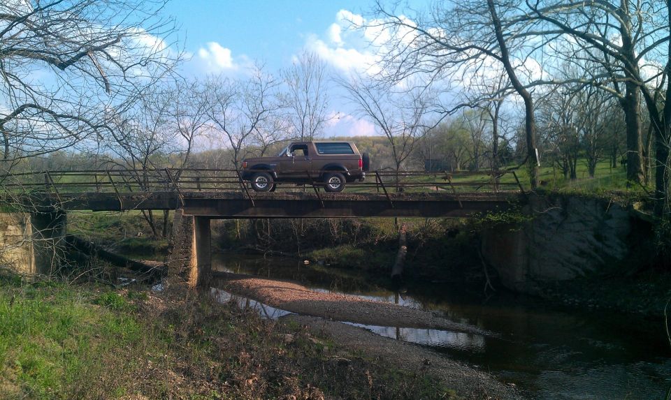Brown 1987 Ford Bronco driving across a rural bridge